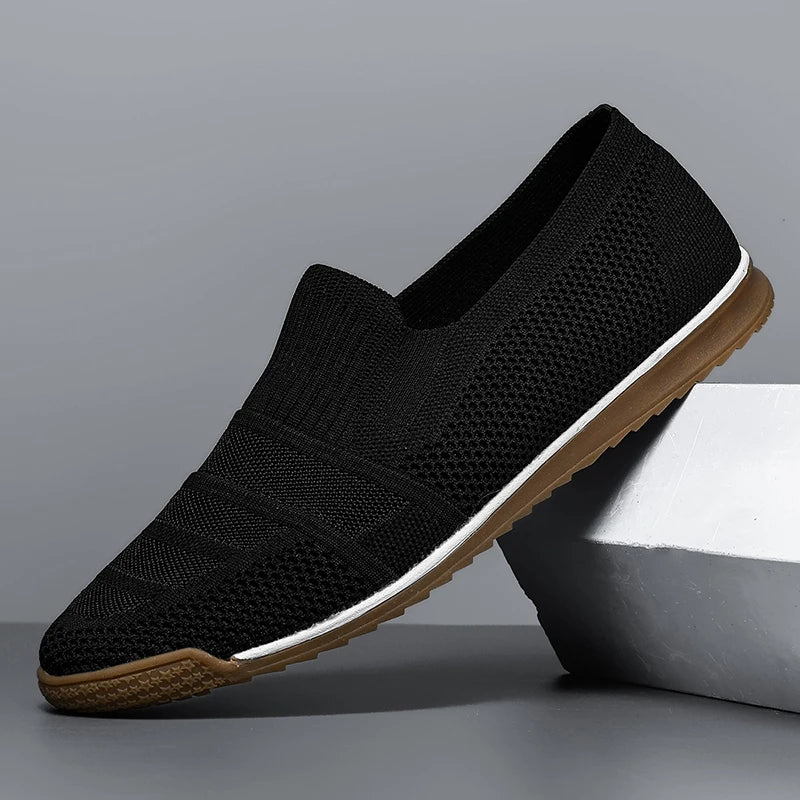 Black slip-on knit men’s casual loafers with a textured upper, canvas lining, white midsole, and brown rubber outsole, displayed at an angle against a gray background with part of the shoe resting on a geometric block.