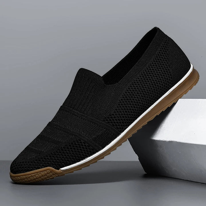 Black slip-on knit men’s casual loafers with a textured upper, canvas lining, white midsole, and brown rubber outsole, displayed at an angle against a gray background with part of the shoe resting on a geometric block.