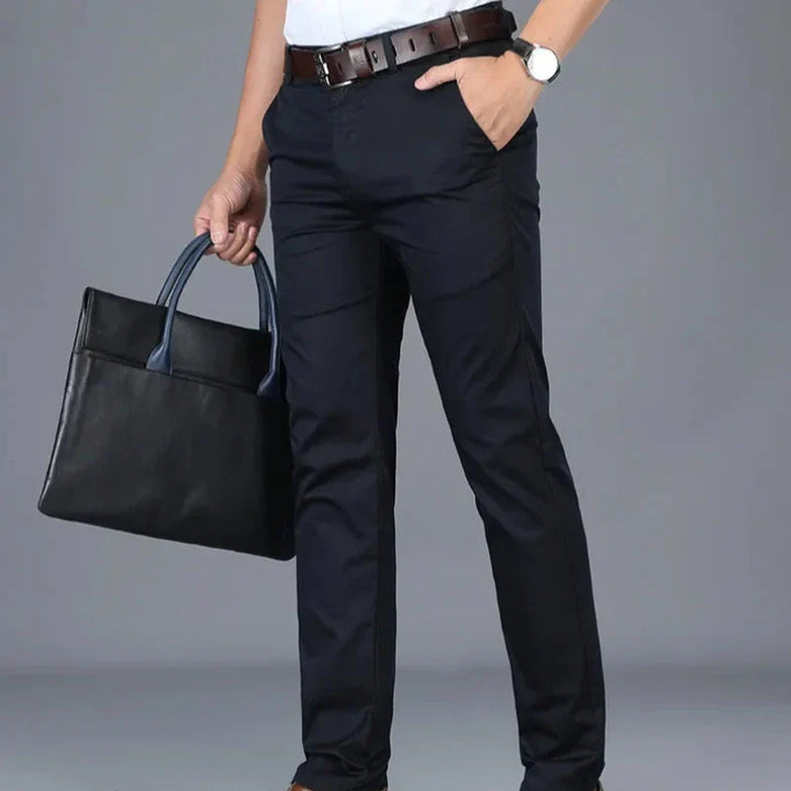 A person dressed in tailored fit dark trousers, a white shirt, and a brown belt stands holding a black briefcase in one hand. The person also wears a wristwatch. The background is plain and gray.