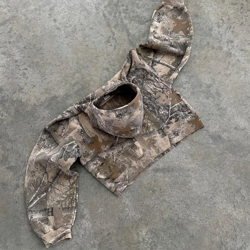 A camo pattern hoodie with long sleeves, crafted from a soft cotton blend, is laid out flat on a textured gray concrete floor. The sleeves are spread wide and the hood is facing up.