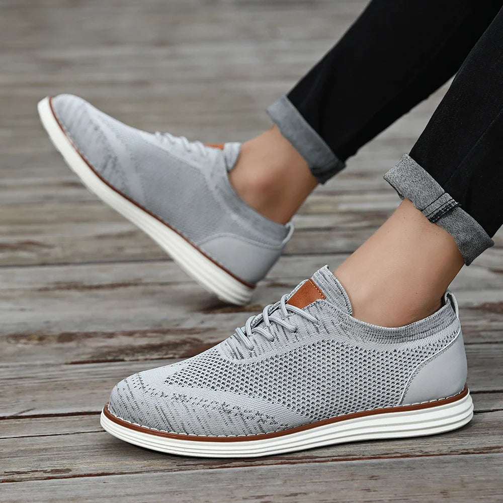 A person wearing breathable knit sneakers in light gray with white soles and brown accents, standing on a wooden surface. The persons black pants are rolled up at the ankles for a casual style.