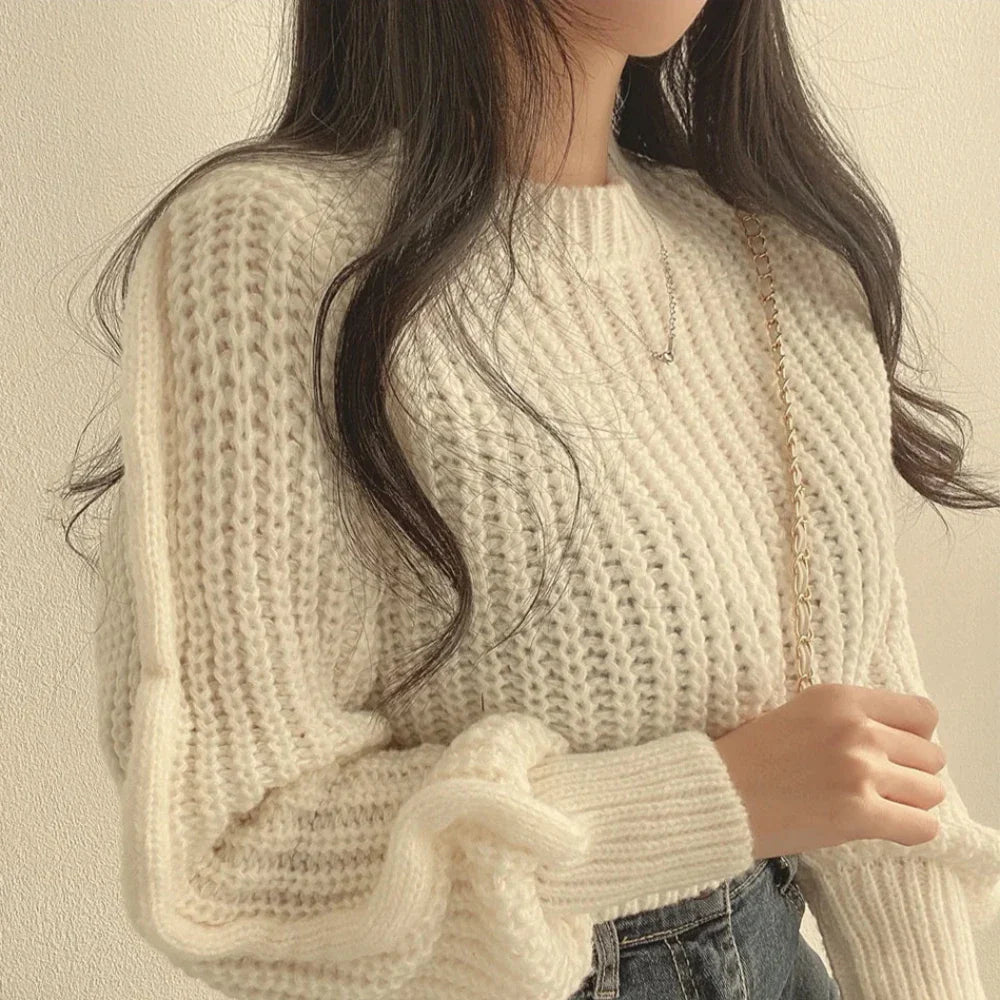 A woman with long, wavy hair wears a chunky white sweater made from soft knit material and holds a gold chain strap over her shoulder. Her face is partially out of frame as she stands against a light-colored wall.