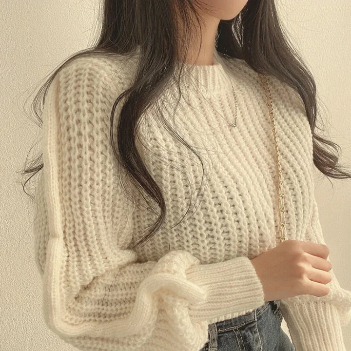 A woman with long, wavy hair wears a chunky white sweater made from soft knit material and holds a gold chain strap over her shoulder. Her face is partially out of frame as she stands against a light-colored wall.