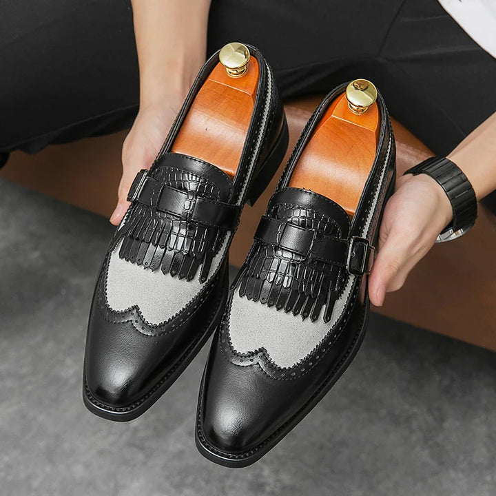 A person holds a pair of stylish black premium leather shoes with gray suede and fringe detailing on the upper, featuring a slip-on design and wooden shoe trees inside.