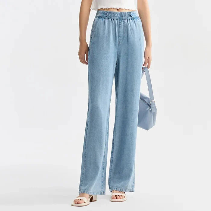 A person wears light blue, high-waisted, wide-leg jeans crafted from breathable denim, a white cropped top, white sandals, and holds a light blue handbag. The background is plain and light-colored.