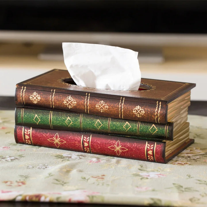A decorative tissue box designed to look like a stack of three vintage books—one red, one green, and one brown—sits on a floral tablecloth, making it perfect as unique home library decor.