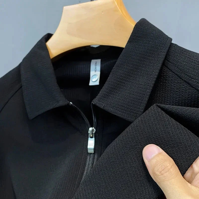 A close-up of a black zip-up collared jacket on a wooden hanger, with a hand holding the textured sleeve to show the fabric details, reminiscent of the smooth finish found in a mens silk polo shirt.