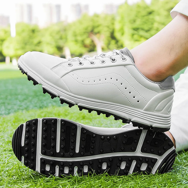 A close-up of a person wearing advanced cleat technology golf footwear, standing on a green grass field, with trees and buildings blurred in the background. One shoe is on the ground while the other foot is lifted, showing the sole.