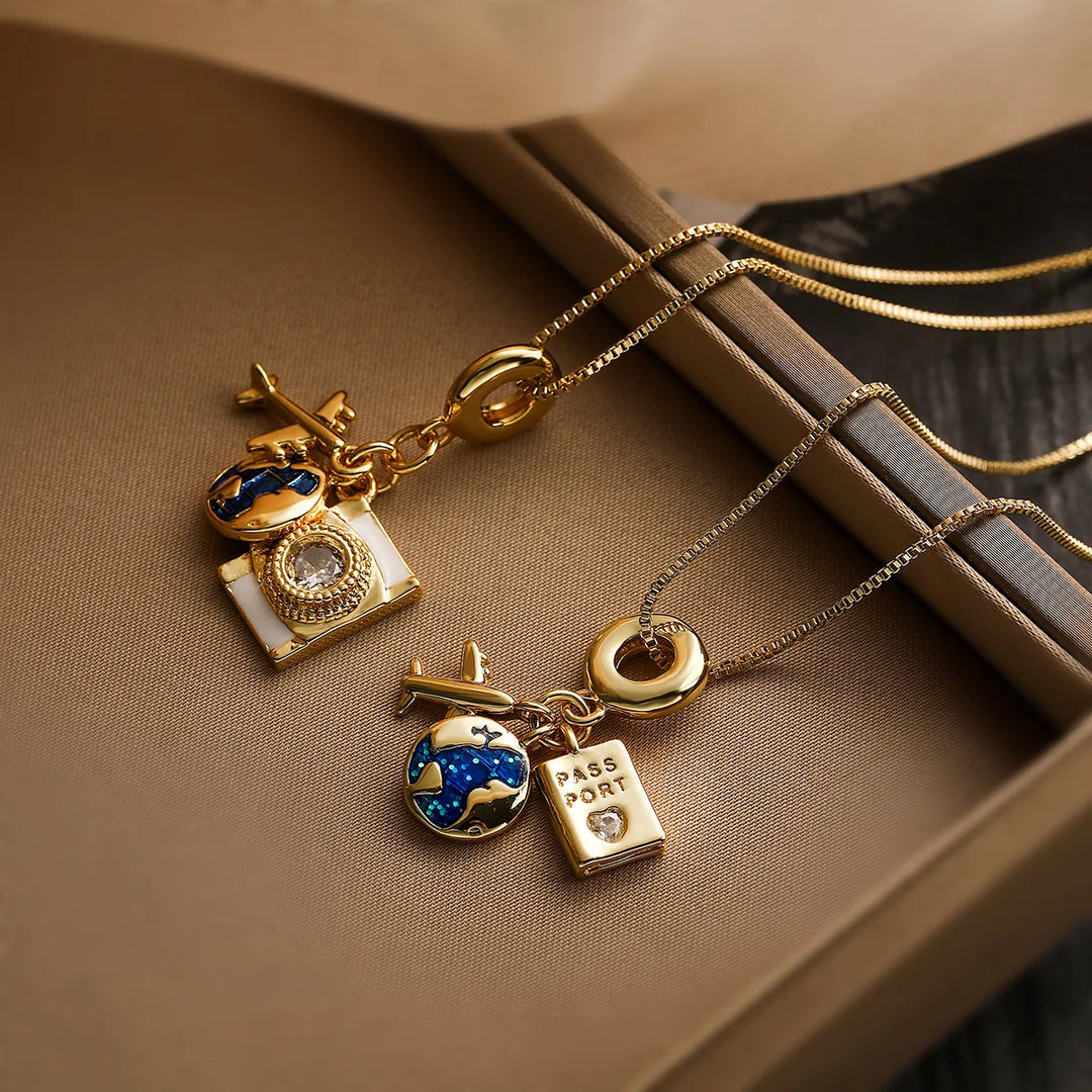 Two gold adventure necklaces on a brown surface, each featuring pendants—a globe, airplane, tiny passport, and jewel—crafted with fine link craftsmanship and a delicate chain, symbolizing travel and discovery.