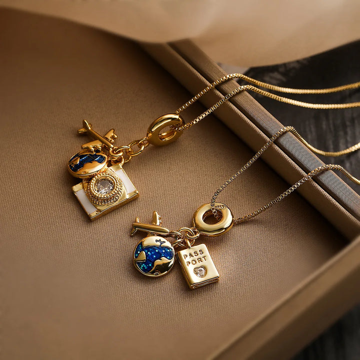 Two gold adventure necklaces on a brown surface, each featuring pendants—a globe, airplane, tiny passport, and jewel—crafted with fine link craftsmanship and a delicate chain, symbolizing travel and discovery.