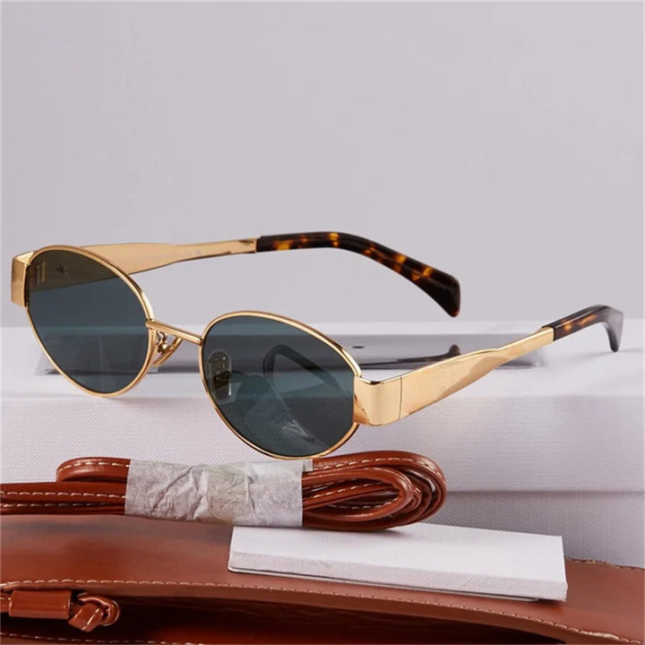 Gold-framed, vintage-inspired sunglasses with oval dark lenses and tortoiseshell arms offer UV400 protection. Displayed on a white surface, they’re paired with a brown leather belt and a white paper-wrapped object in the foreground.