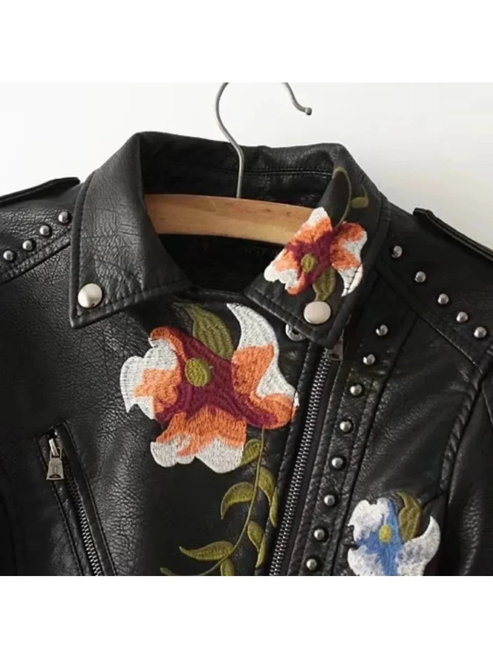 A black PU Leather Jacket with silver studs and zippers, featuring colorful embroidered flowers on the collar and chest—an edgy Embroidered Moto Jacket hanging on a wooden hanger.