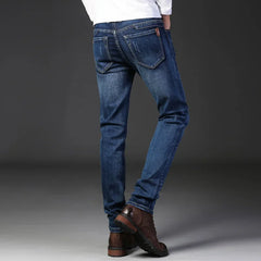 A person wearing slim-fit jeans made from durable fabric and brown leather shoes stands with their back to the camera against a dark, plain background. Only their legs and lower torso are visible.