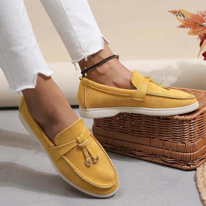 A person wearing mustard yellow chic moccasins with white soles and white frayed-hem jeans, resting their feet near a woven basket and autumn-themed decor.