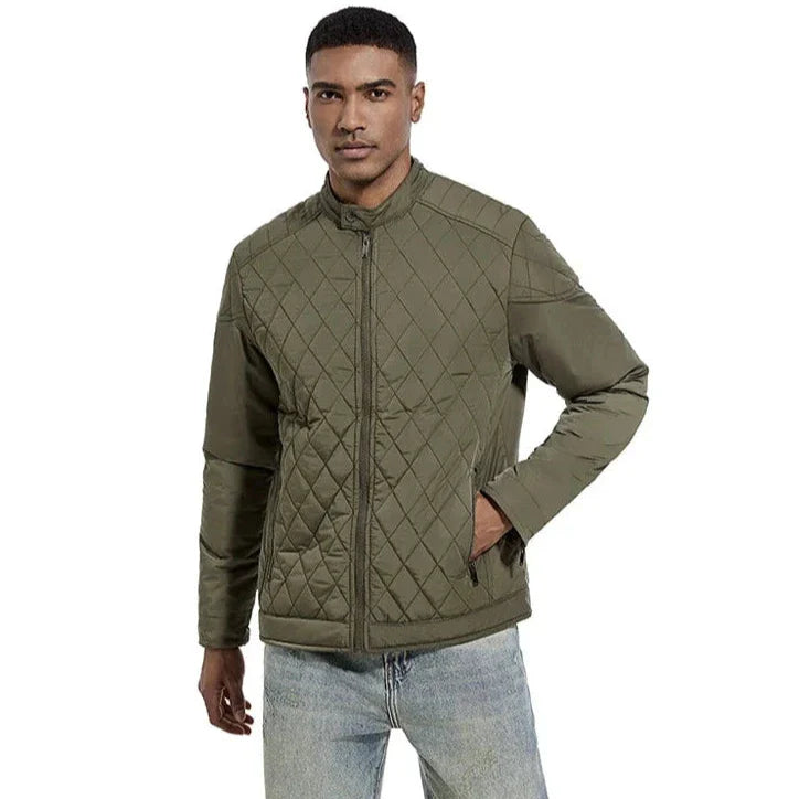 A man wearing a wind-resistant, quilted olive green slim-fit jacket with front zipper and light blue jeans stands against a plain white background, looking forward with one hand in his pocket.
