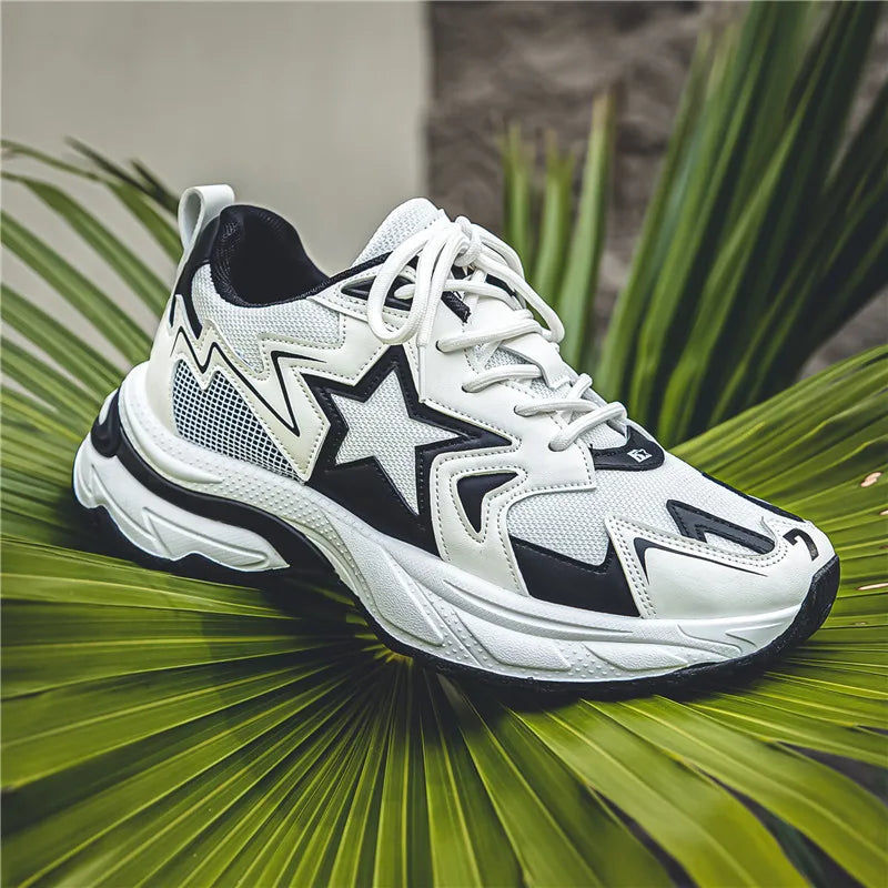 A white athletic sneaker with black star and zigzag patterns, featuring a breathable mesh upper, rests on large green palm leaves against a blurred gray background.