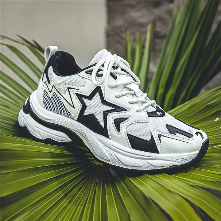 A white athletic sneaker with black star and zigzag patterns, featuring a breathable mesh upper, rests on large green palm leaves against a blurred gray background.