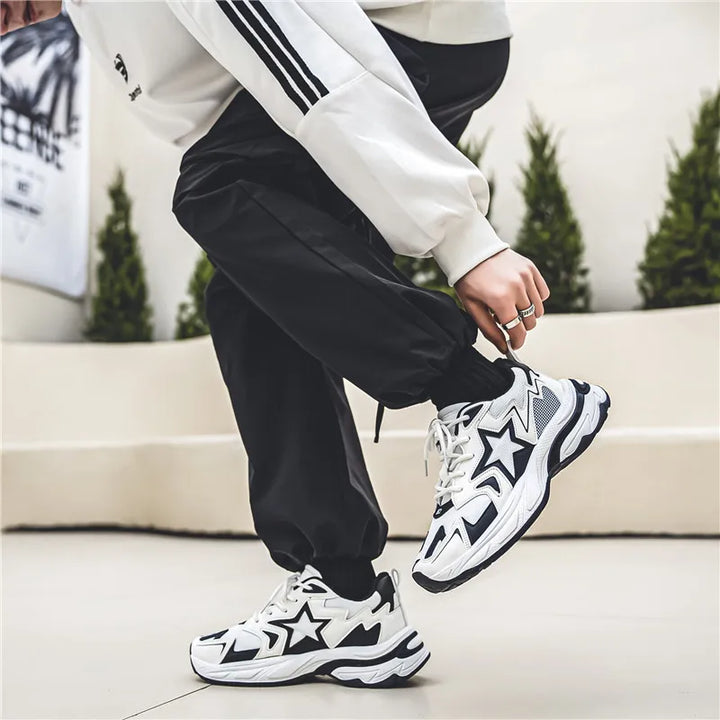 A person wearing black joggers, a white sweatshirt with black stripes, and white sneakers featuring a durable rubber outsole with black star designs is crouching outdoors on a light-colored surface. Green shrubs are in the background.