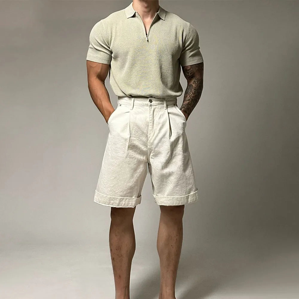 A man stands with hands in pockets, wearing a light beige quarter zip polo and high-waisted, pleated off-white shorts. The background is plain and neutral. His face is not visible.