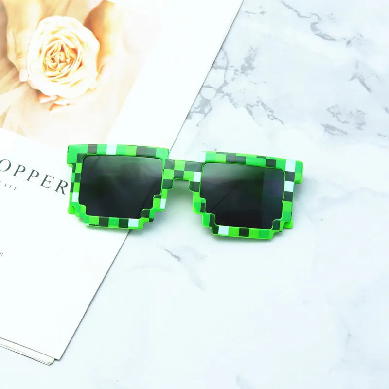 Green and black pixelated sunglasses with square frames, featuring UV400 protection, rest on a light grey marble surface next to an open magazine displaying a close-up of a pale peach rose.