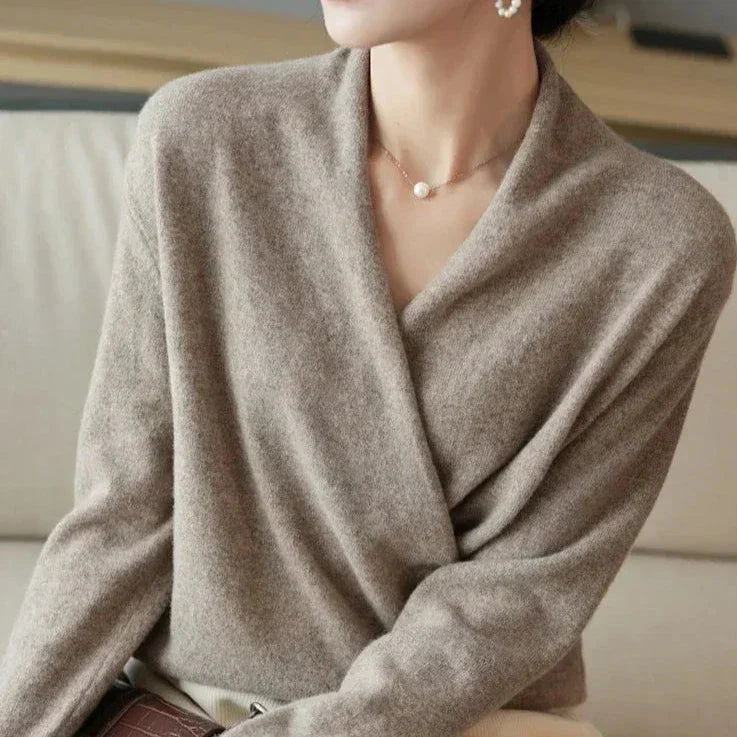 A woman wearing a soft brown cashmere wrap sweater, a simple pearl necklace, and pearl earrings sits on a light-colored sofa. Her face is partially out of frame and her hair is pulled back.