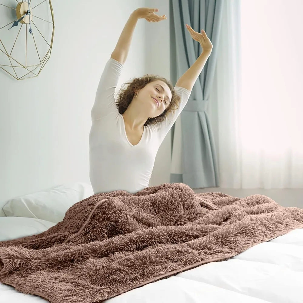 A woman sits up in bed stretching with her arms raised, smiling. She is covered with a soft, fluffy Faux Fur Weighted Blanket, and sunlight streams through a curtained window behind her.