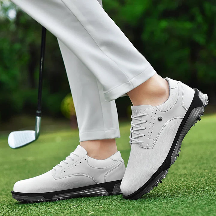 Person wearing white golf shoes with a breathable mesh lining and white pants stands on green grass, holding a golf club. The blurred greenery backdrop highlights the shoes design, texture, and comfort.