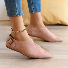 A person wearing light pink pointed toe shoes with a side buckle and gold chain anklet, featuring a cushioned insole, paired with frayed hem blue skinny jeans, standing on a wooden floor.