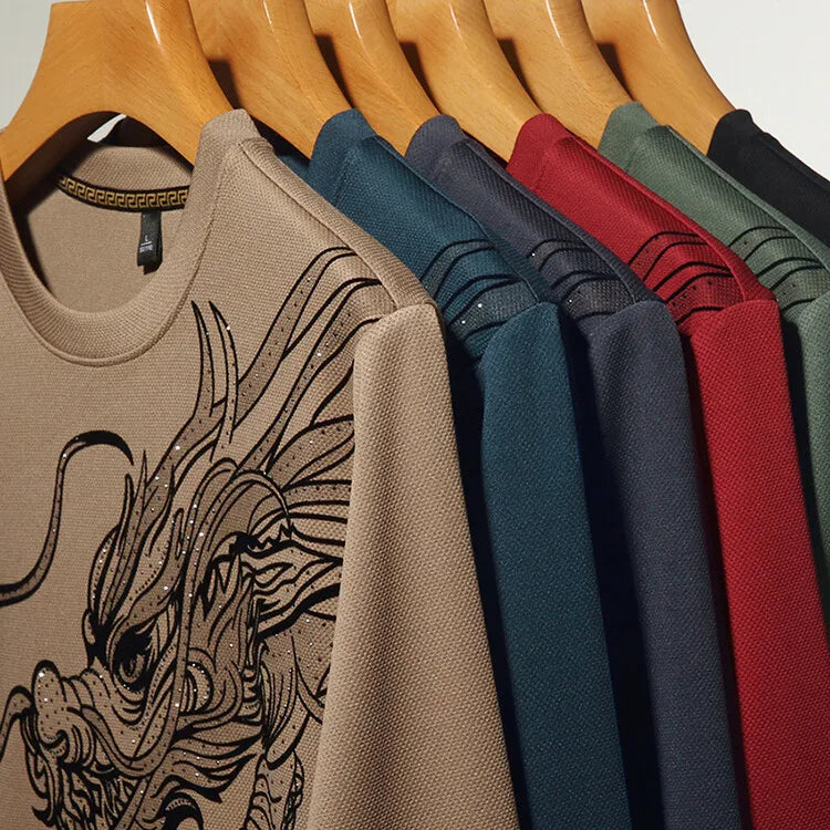 Six mens crewneck shirts on wooden hangers, each a different color—beige, blue, navy, red, green, and gray. The beige ultra-soft cotton crewneck in front features a detailed black dragon graphic on the chest.