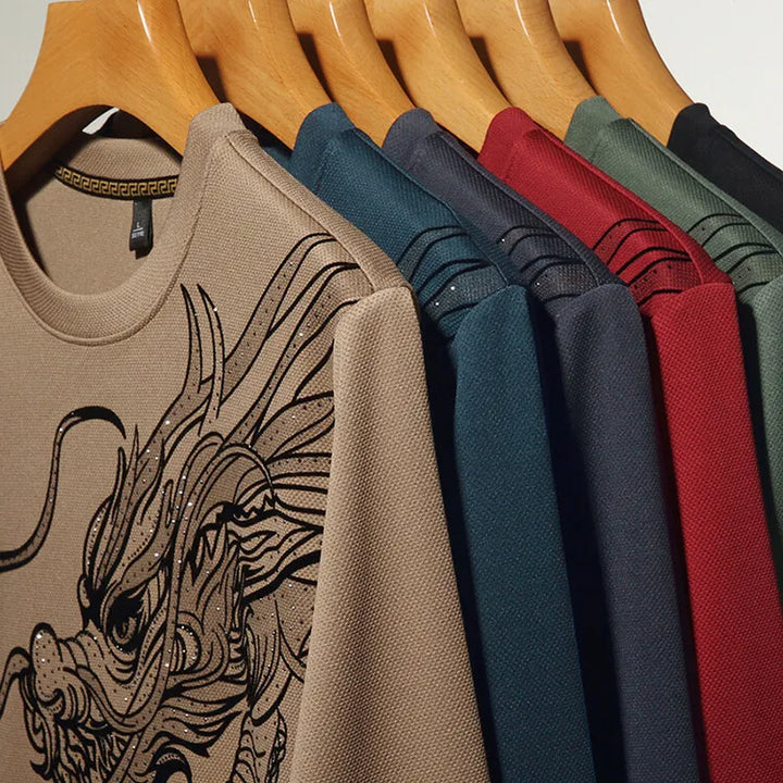 Six mens crewneck shirts on wooden hangers, each a different color—beige, blue, navy, red, green, and gray. The beige ultra-soft cotton crewneck in front features a detailed black dragon graphic on the chest.