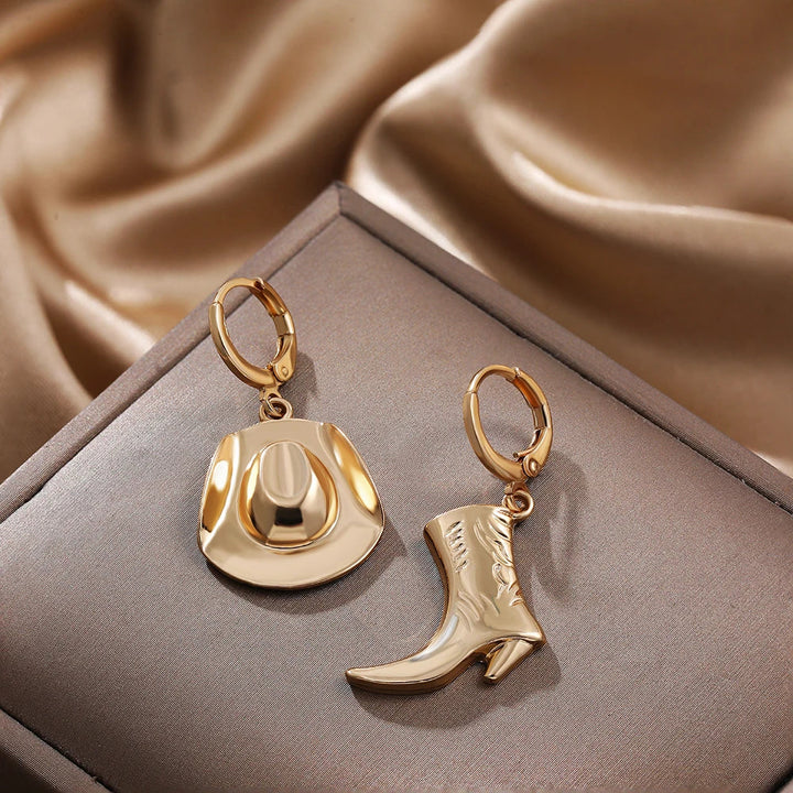A pair of gold hoop earrings, one with a cowboy hat charm and the other with a cowboy boot charm, displayed on a brown jewelry box—perfect western jewelry for those who love unique cowgirl accessories.