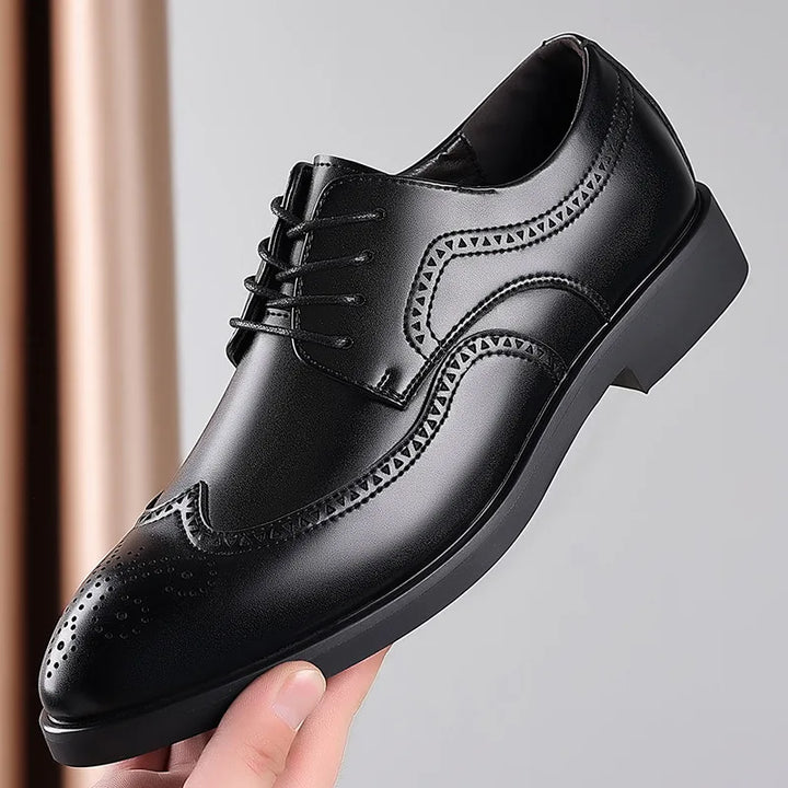 A hand holds up a polished black premium leather dress shoe with brogue detailing, decorative perforations, and stitching, featuring a lace-up design and a slightly raised heel against a neutral background.