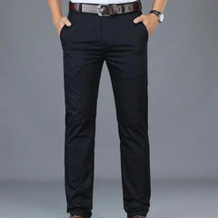 A person wearing dark blue dress pants featuring a tailored fit and hands in pockets, a brown belt, a gray shirt tucked in, and a wristwatch, standing against a plain gray background.