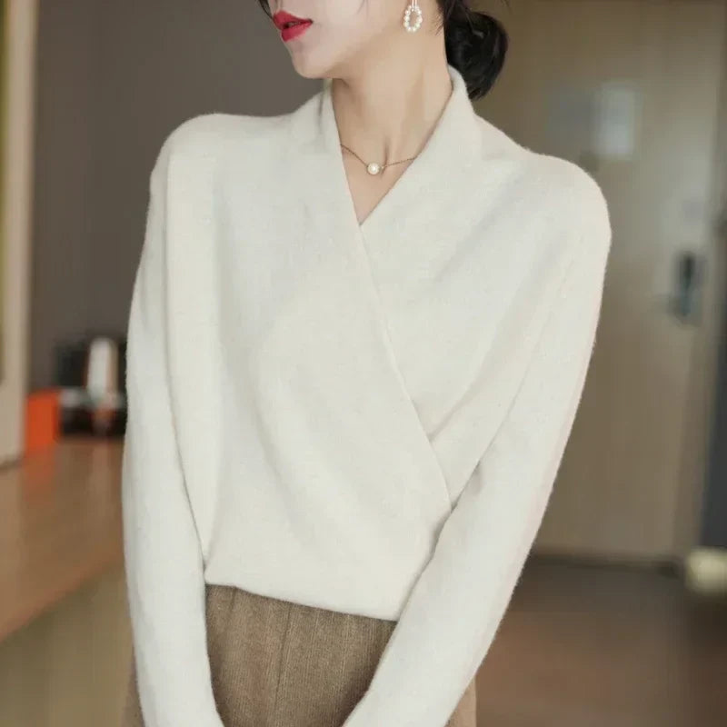 Woman wearing a white wrap-style sweater crafted from luxurious fabric, brown pants, drop earrings, and a delicate necklace, standing indoors with a blurred background. Her face is partially turned to the side.