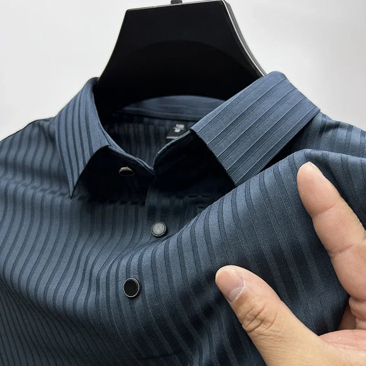 A close-up of a dark blue, vertically striped dress shirt on a hanger, with a hand holding the luxurious silk fabric to show its texture and softness. The shirt has black buttons and a pointed collar.