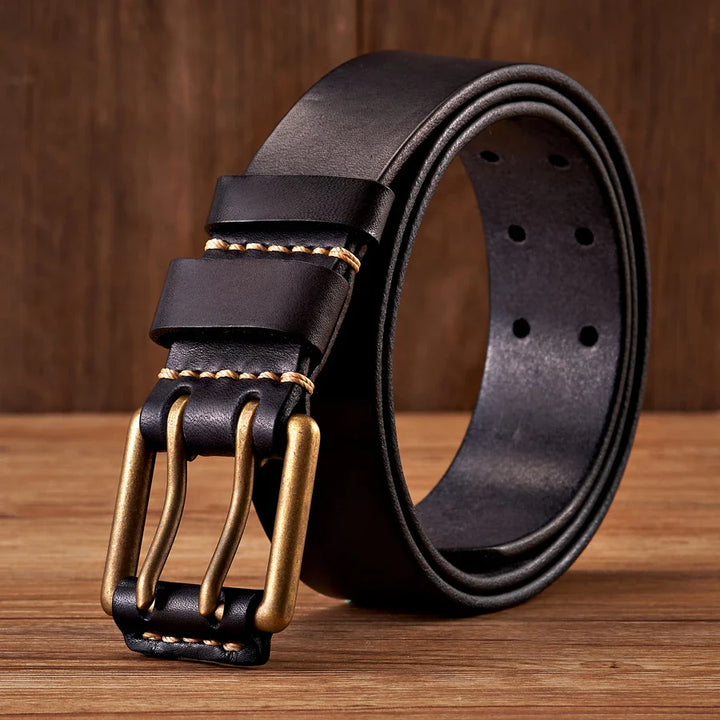 A durable cowhide leather belt with a brass buckle and tan stitching, displayed upright on a wooden surface against a wood-paneled background.