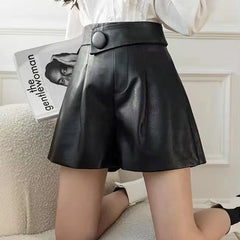 A woman wears high waist black calfskin leather shorts with a large button detail, sitting on a cream textured chair while holding a magazine with a black and white cover.