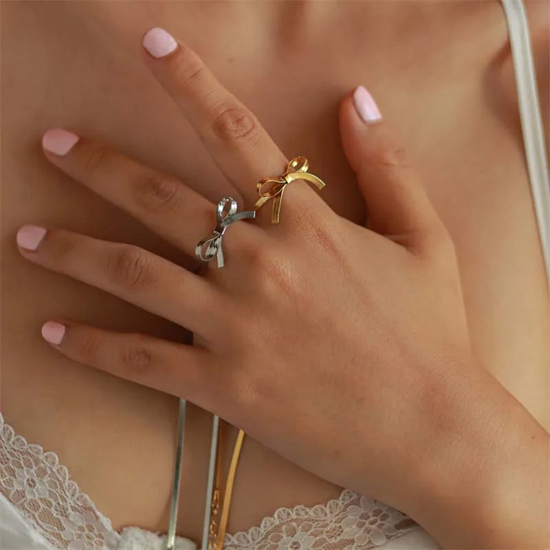 A close-up of a womans hand with light pink nail polish, wearing delicate gold and silver bow-shaped rings—perfect for pairing with minimalist jewelry like a dainty bow necklace. She is dressed in a white lace-trimmed top.