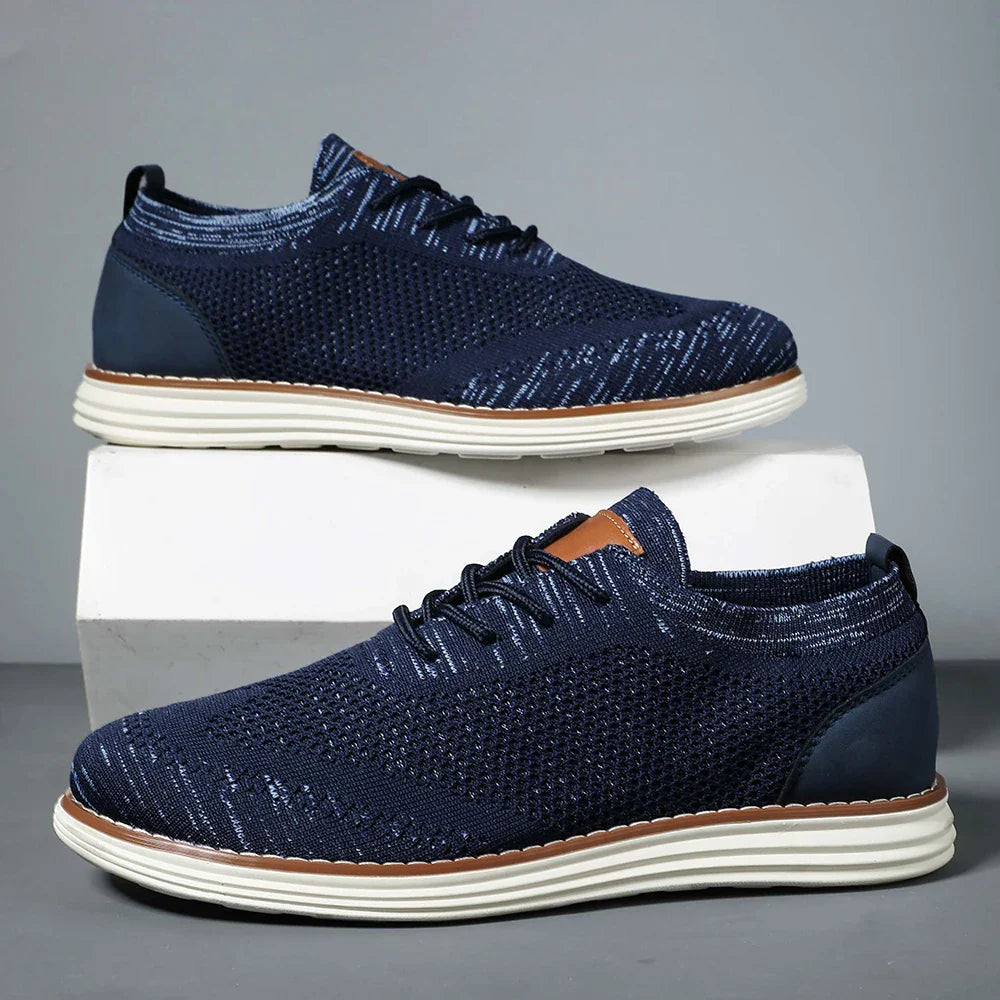 A pair of navy blue breathable knit sneakers with white soles and black laces, displayed against a gray background. One shoe is elevated on a white rectangular block, highlighting their casual style and the side profile of both shoes.