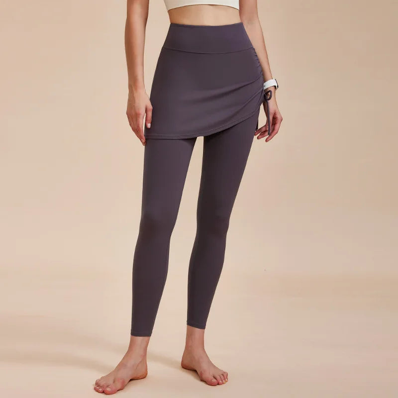 A person models high-waisted leggings with skirt in dark gray, perfect for an autumn wardrobe, standing barefoot against a plain beige background. Their face is not visible.