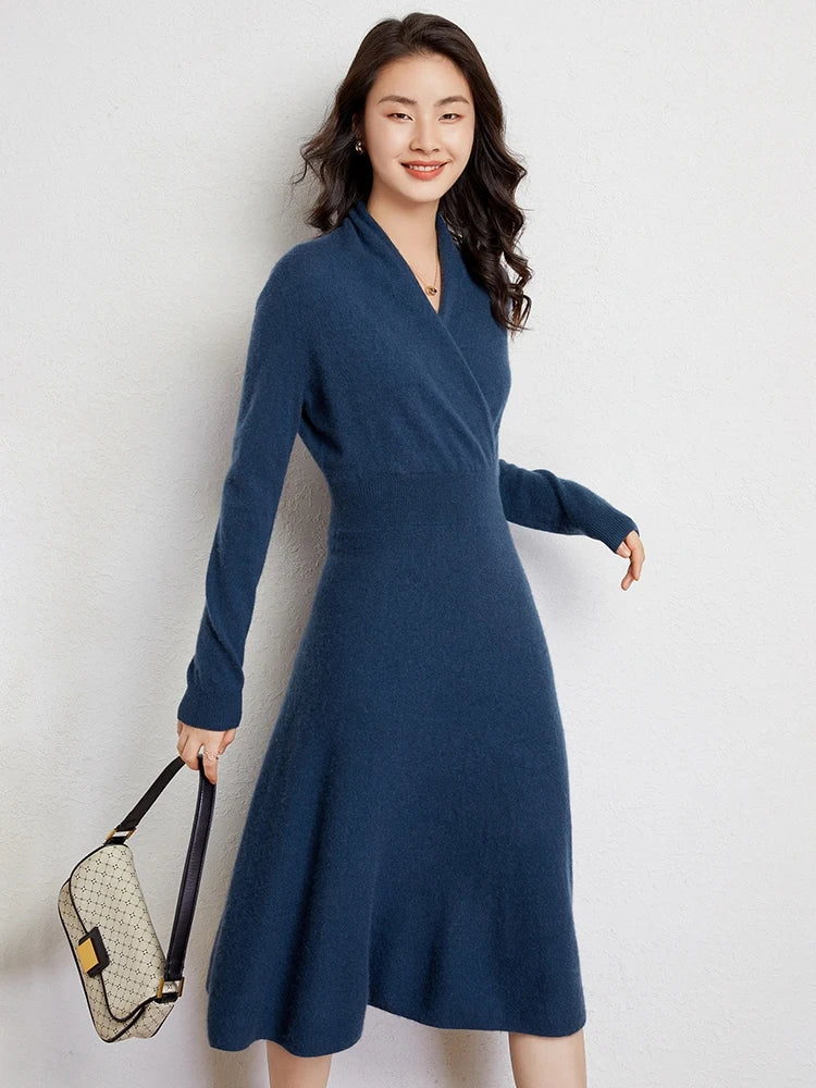 A woman with long wavy hair smiles while wearing a knee-length blue Matilda Wrap Dress featuring a V-neck wrap style. She stands against a plain light wall, holding a beige handbag with a dark strap in her left hand.