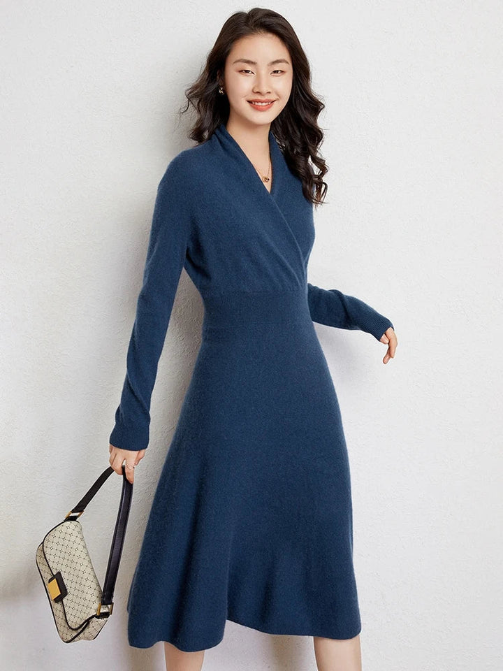 A woman with long wavy hair smiles while wearing a knee-length blue Matilda Wrap Dress featuring a V-neck wrap style. She stands against a plain light wall, holding a beige handbag with a dark strap in her left hand.