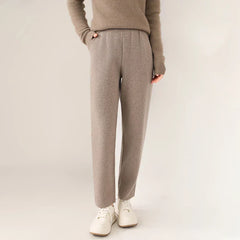 A person wears a taupe sweater and matching slim tailored leg trousers with white sneakers, standing against a plain light background with hands in pockets.