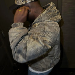 A person wearing a cotton blend camo pattern jacket and matching cap stands indoors, turning their head and touching their face, partially obscuring it from view.