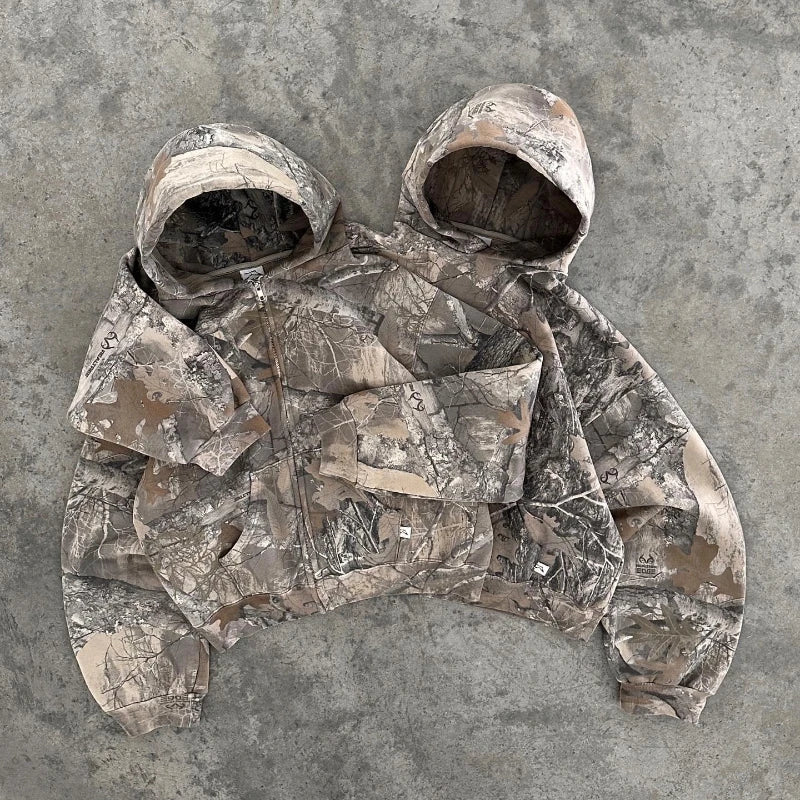 Two camo pattern hooded jackets are sewn together side by side, sharing a middle sleeve, and laid flat on a textured concrete surface.