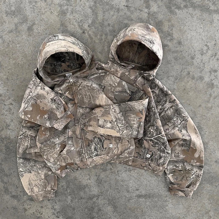 Two camo pattern hooded jackets are sewn together side by side, sharing a middle sleeve, and laid flat on a textured concrete surface.