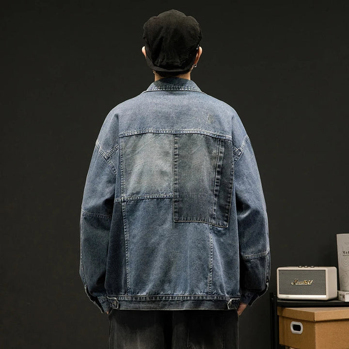 A person in an oversized fit blue denim jacket and a black cap stands with their back to the camera. The dark background features a small speaker and some papers on a wooden surface to the right.