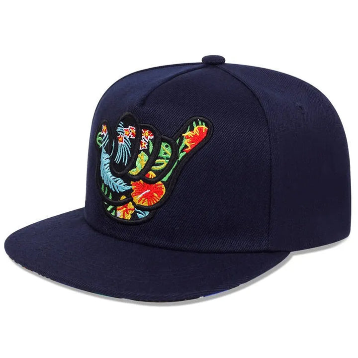 A navy blue Hang Loose Snapback featuring an embroidered shaka hand sign filled with a colorful tropical floral pattern on the front panel—perfect for that Hawaiian-inspired cap vibe.