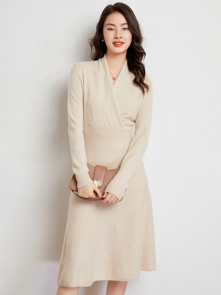 A woman with long, wavy dark hair wears a light beige Matilda Wrap Dress featuring a V-neck wrap style and holds a taupe clutch purse, standing against a plain light-colored wall and smiling softly.
