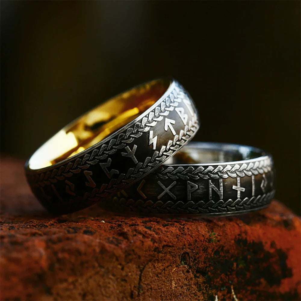 Two Viking Rune Rings with engraved runic symbols rest on a reddish-brown textured surface. Crafted as stainless steel rings, their inner side gleams gold while the outer surfaces display intricate patterns—ideal as couple's amulet rings.